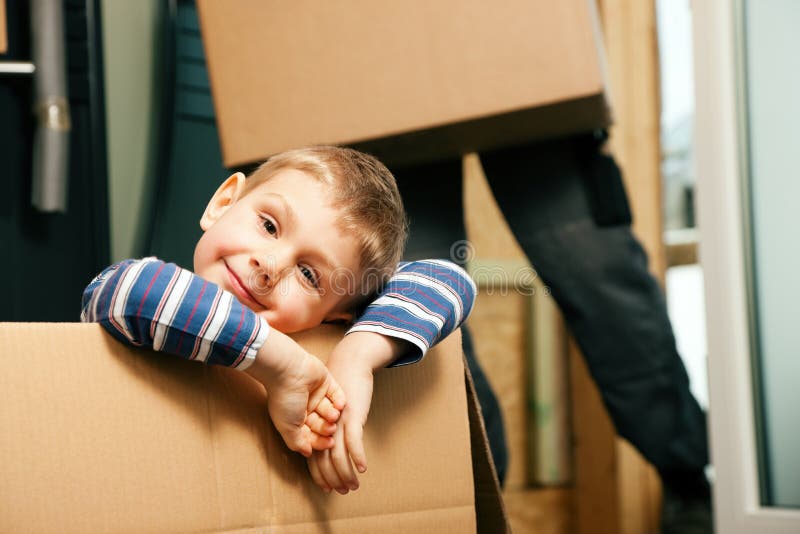 Family moving in their new home. The son is sitting inside a moving box. In the background the father - or a mover (only legs to be seen) is carrying boxes inside the building. Family moving in their new home. The son is sitting inside a moving box. In the background the father - or a mover (only legs to be seen) is carrying boxes inside the building