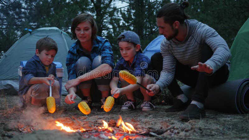 Família que prepara o milho amarelo fritado na noite da noite no resto na floresta, piquenique fora, família americana