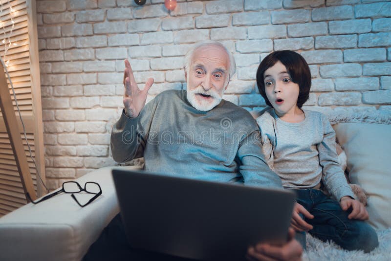 Farfadern och sonsonen sitter hållande ögonen på intressant film på natten hemma arkivbilder Farfadern och sonsonen sitter hållande ögonen på intressant film på natten hemma arkivbilder