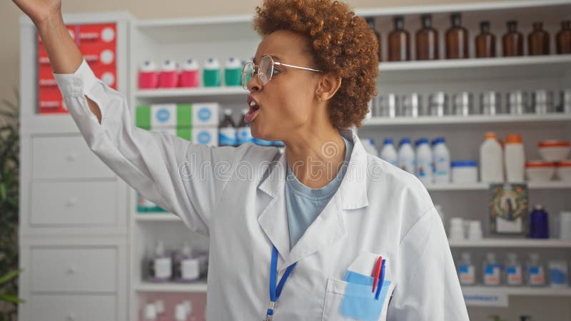 Farmacéutica afroamericana gritando en una concurrida farmacia de hospital llevando gafas y abrigo de laboratorio imagen de archivo libre de regalías