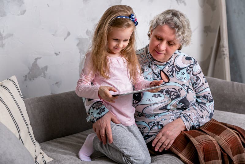 Farmodern och hennes lilla sondotter h?ller ?gonen p? filmer tillsammans och spelar p? apparaten, medan sitta p? soffan royaltyfri fotografi Farmodern och hennes lilla sondotter h?ller ?gonen p? filmer tillsammans och spelar p? apparaten, medan sitta p? soffan royaltyfri fotografi