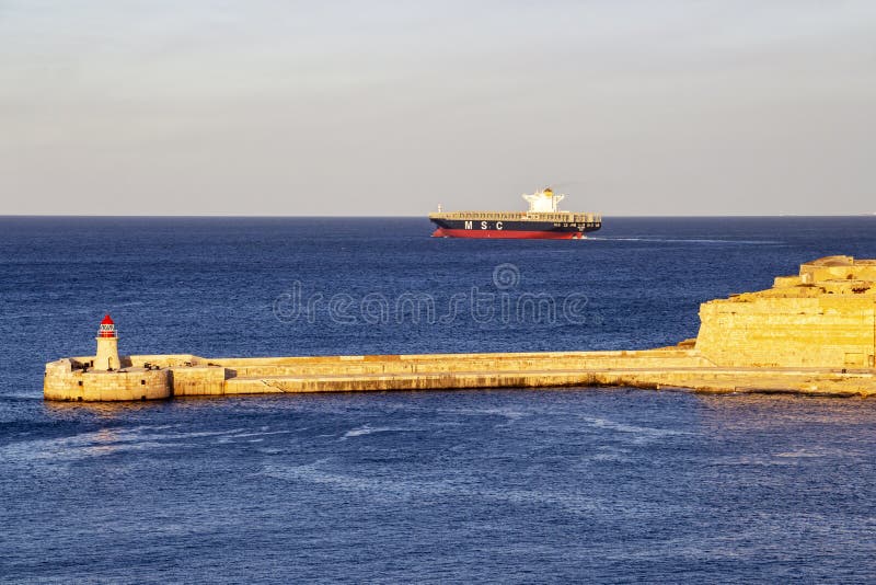 Farol do quebra-mar de Ricasoli no porto grande, Malta com um navio de recipiente imagens de stock royalty free Farol do quebra-mar de Ricasoli no porto grande, Malta com um navio de recipiente imagens de stock royalty free