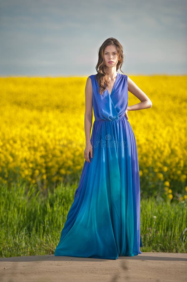 Fashion beautiful young woman in blue dress posing outdoor with cloudy dramatic sky in background. Attractive long hair brunette