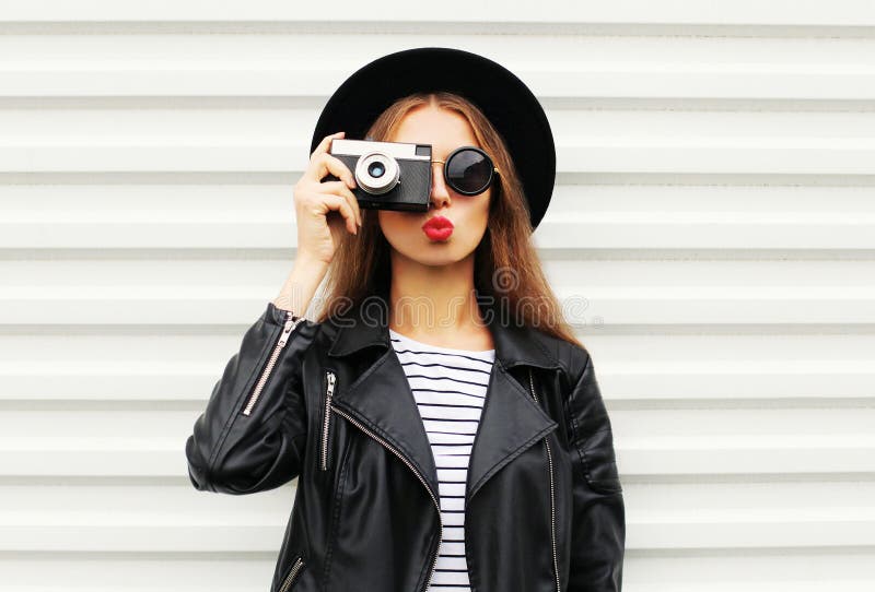 Fashion look, pretty cool young woman model with retro film camera wearing elegant black hat, leather rock jacket over white