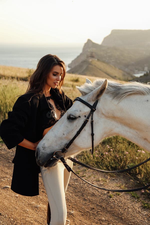 Beautiful woman with dark hair in elegant clothes posing with white horse at  landscape with mountains and fields