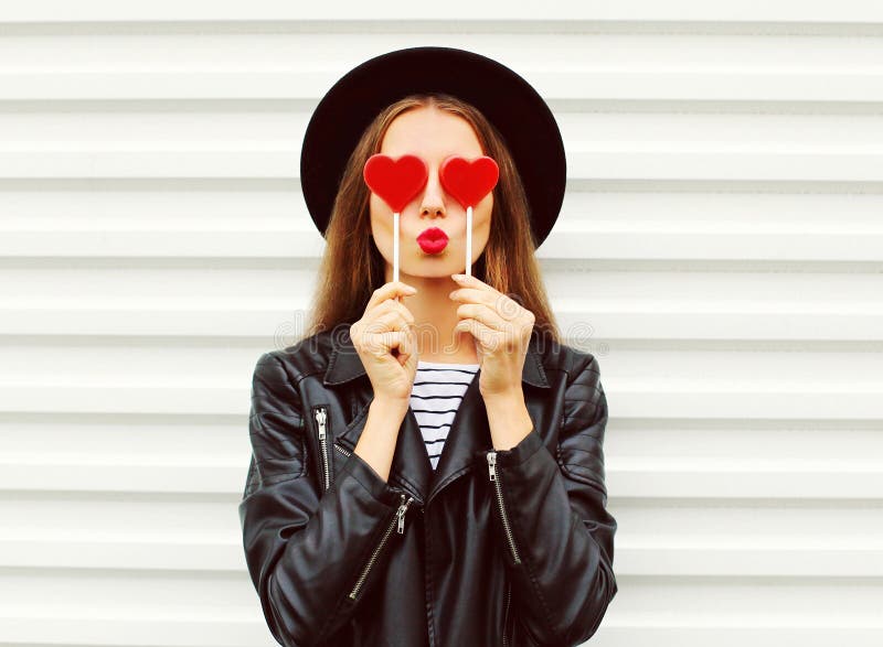 Fashion portrait pretty sweet young woman with red lips making air kiss with lollipop heart wearing black hat leather jacket