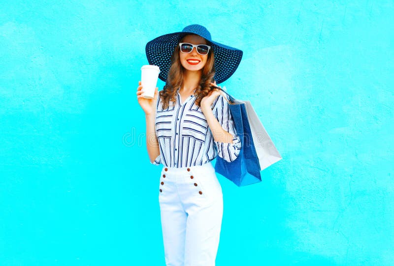 Fashion pretty young smiling woman with coffee cup wearing a shopping bags, straw hat, white pants over colorful blue background