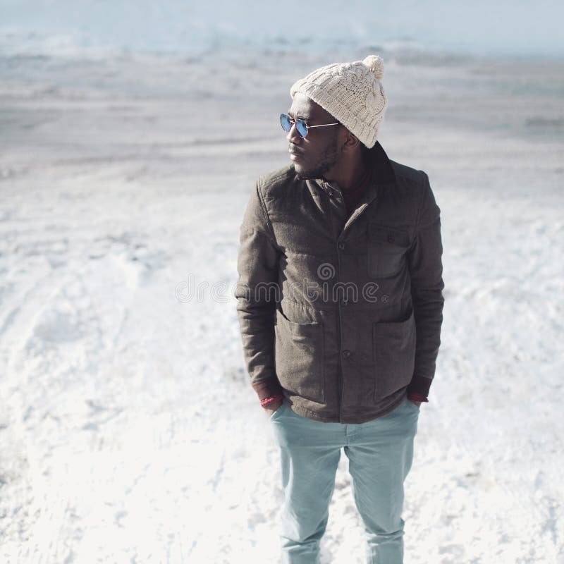 Fashion stylish young african man wearing a sunglasses, knitted hat and jacket in winter day over snow