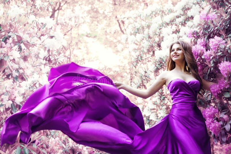 Fashion Woman Pink Dress in Spring Flower Garden. Beauty Model Girl in Purple Silk Gown Fluttering on Wind. Floral Pink Fantasy