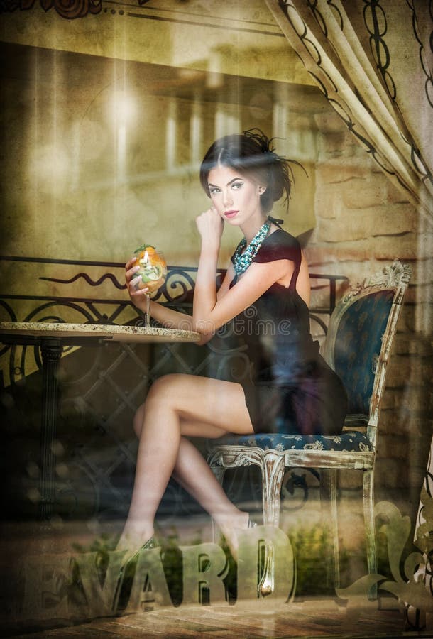 Fashionable attractive young woman in black dress sitting in restaurant, beyond the window. Beautiful brunette posing in window