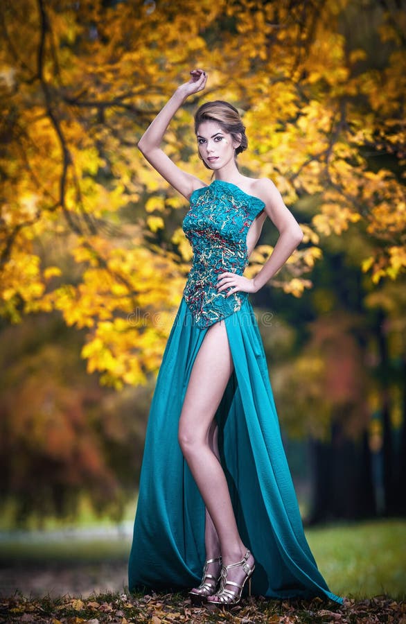 Fashionable beautiful young woman in blue dress posing outdoor rusty forest in background. Attractive girl with elegant dress posing in autumnal park. Luxurious elegant lady in blue, autumnal concept