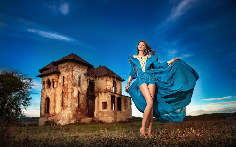 Fashionable beautiful young woman in long blue dress posing with old castle and cloudy dramatic sky in background