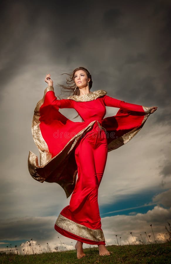 Fashionable beautiful young woman in red long dress posing outdoor with cloudy dramatic sky in background