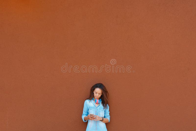 Fashionable cool girl in headphones listening to music on the phone against the background of the city wall. Concept of urban style, youth and trends, feminism, emancipation, womens rights