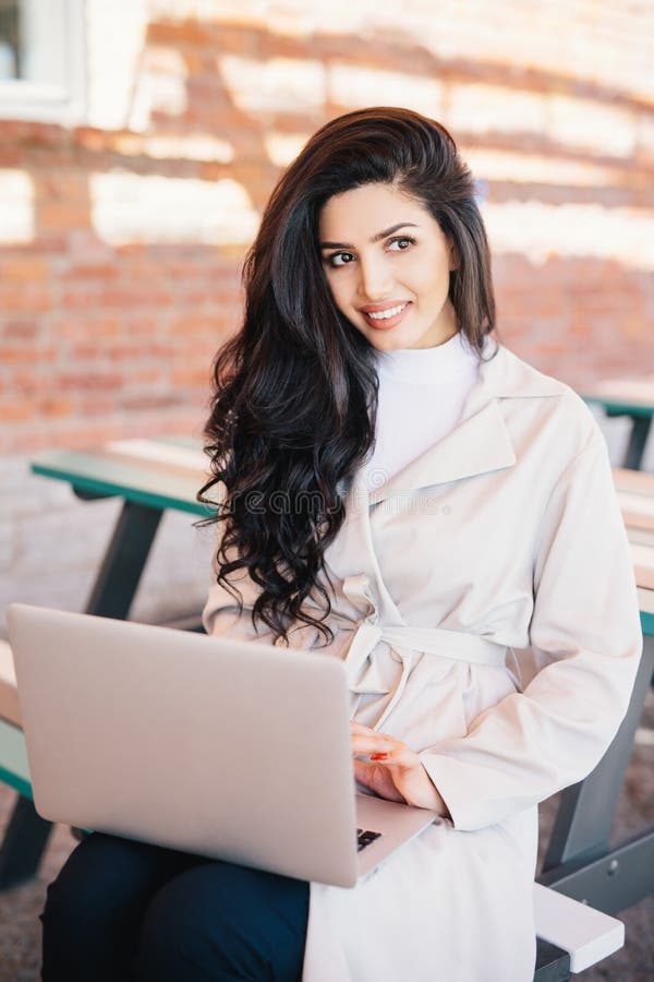 Fashionable female model in elegant white coat sitting at bench holding laptop computer surfing social networks. Professional bussinesswoman sitting at outdoor cafe working with modern computer