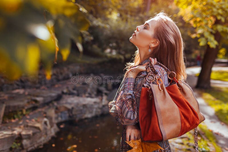 Fashionable woman holds brown purse in autumn park. Stylish girl wears trendy accessories and clothes walking outdoors