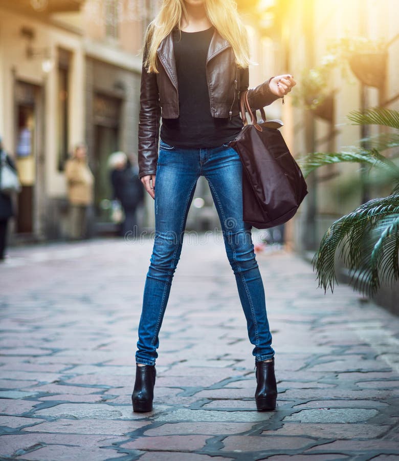 Fashionable young blonde girl with long legs wearing blue jeans, leather brown coat and holding a bag walking and shopping on city