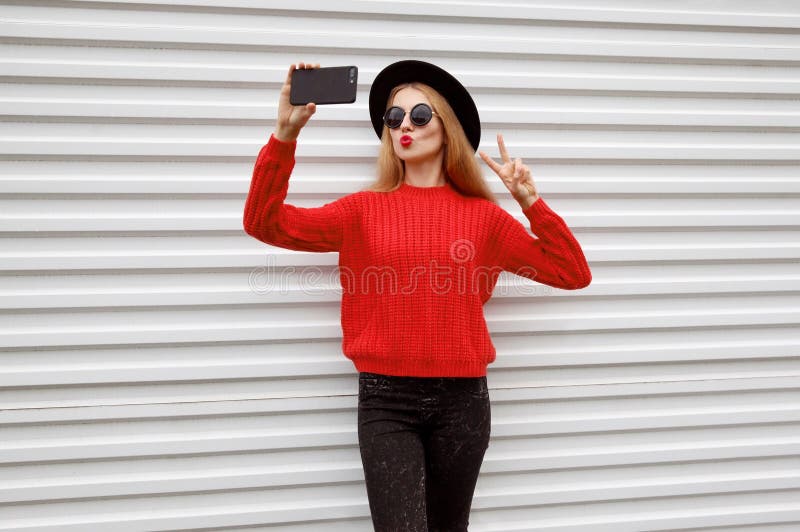 Fashionable young woman blowing red lips sends sweet air kiss taking selfie picture by phone in knitted sweater stock photos Fashionable young woman blowing red lips sends sweet air kiss taking selfie picture by phone in knitted sweater stock photos