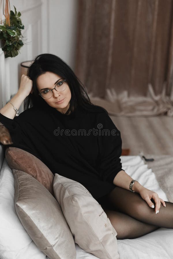 A fashionable young woman with perfect dark hair in an elegant black dress and round glasses posing on the bed
