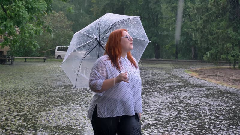 Fat ginger girl with glasses is walking in park under rain, watching up, holding umbrella