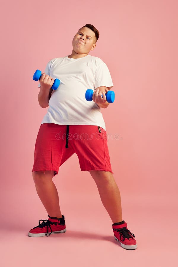 Fat young boy wearing white tee and red shorts doing sport exercises isolated on pink background. Sport, vacation