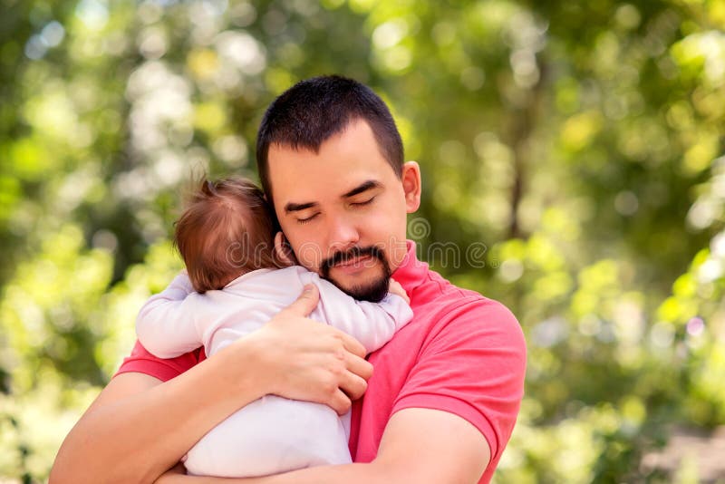 Father gently holding in arms infant daughter in pink jumpers outdoors in warm sunny or early autumn day. Baby is hugging dad