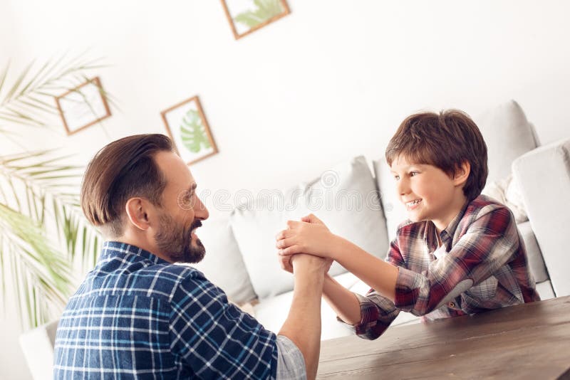 Father and little son at home sitting at table arm wrestling boy holding dad`s hand smiling cheerful royalty free stock images Father and little son at home sitting at table arm wrestling boy holding dad`s hand smiling cheerful royalty free stock images