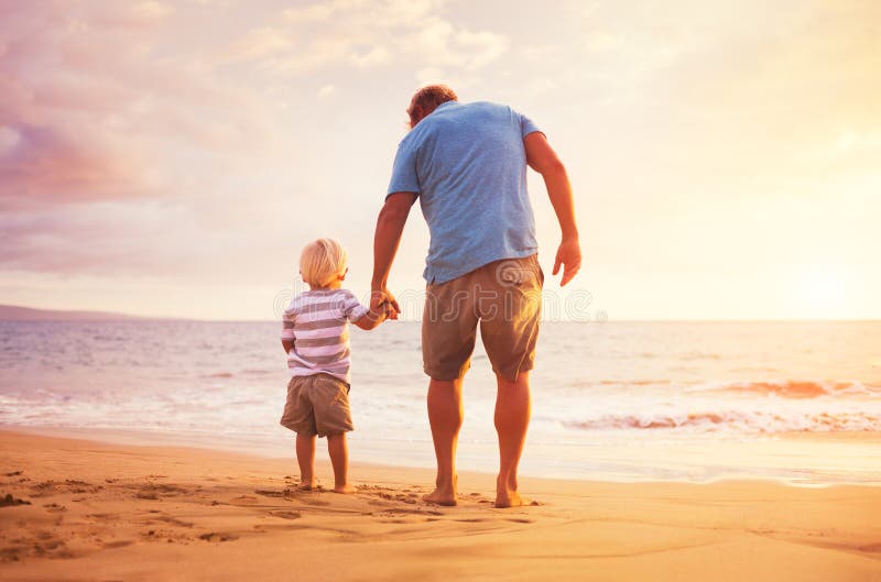 Otec a syn, ktorý stojí na brehu mora držanie ruky pri západe slnka.