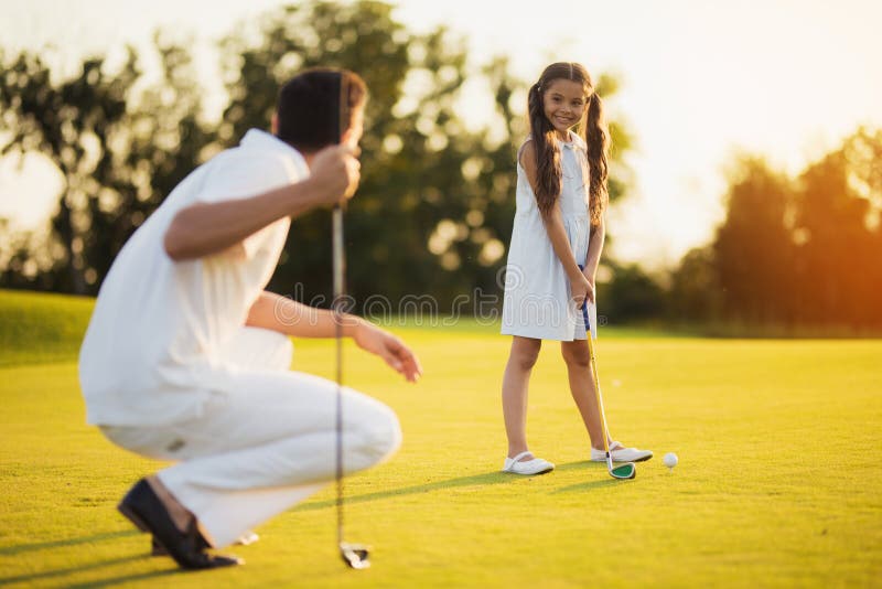 The father squats with a golf club in his hand and looks at his daughter, who looks at him and prepares to hit the ball stock image The father squats with a golf club in his hand and looks at his daughter, who looks at him and prepares to hit the ball stock image