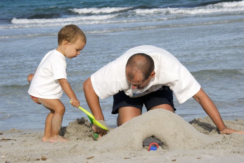Otec učí malé kavkazské chlapeček k budování hrad z písku tváří v tvář moři, v letní den.