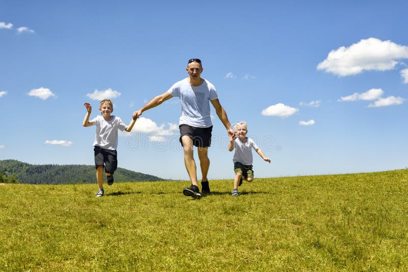 Father with two young children running hand in hand on the green field on a background of blue sky and clouds. Fatherhood and stock image Father with two young children running hand in hand on the green field on a background of blue sky and clouds. Fatherhood and stock image