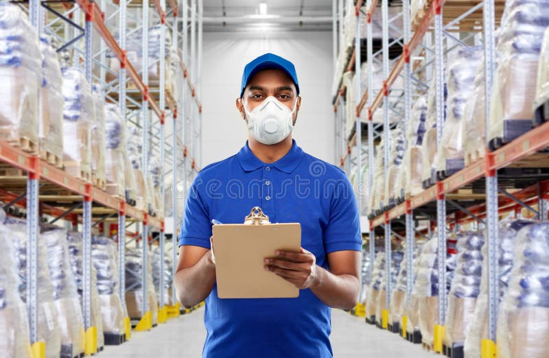 Health, safety and pandemic concept - happy indian delivery man in blue uniform wearing face protective mask or respirator for protection from virus with clipboard over warehouse background. Health, safety and pandemic concept - happy indian delivery man in blue uniform wearing face protective mask or respirator for protection from virus with clipboard over warehouse background