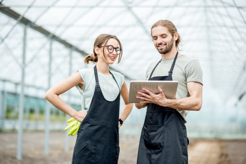 Couple of farmers in uniform working with digital tablet standing in the greenhouse with cultivated soil ready for planting. Couple of farmers in uniform working with digital tablet standing in the greenhouse with cultivated soil ready for planting