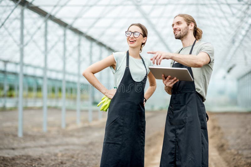 Couple of farmers in uniform working with digital tablet standing in the greenhouse with cultivated soil ready for planting. Couple of farmers in uniform working with digital tablet standing in the greenhouse with cultivated soil ready for planting