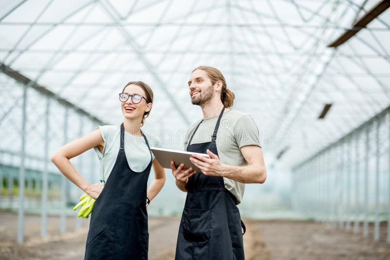 Couple of farmers in uniform working with digital tablet standing in the greenhouse with cultivated soil ready for planting. Couple of farmers in uniform working with digital tablet standing in the greenhouse with cultivated soil ready for planting