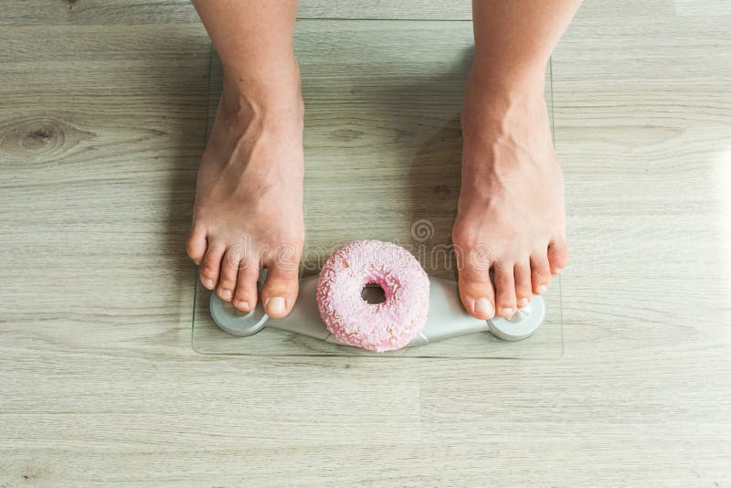 Faça dieta o conceito Close-up dos pés do ` s da mulher na escala de peso com filhós Conceito dos doces, da comida lixo insalubre fotografia de stock royalty free Faça dieta o conceito Close-up dos pés do ` s da mulher na escala de peso com filhós Conceito dos doces, da comida lixo insalubre fotografia de stock royalty free