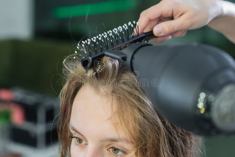 Cabeleireiro secando cabelos loiros à mão com secador de cabelo e escova  redonda em um salão de beleza.