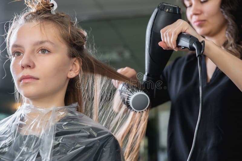 Cabeleireiro secando cabelos loiros à mão com secador de cabelo e escova  redonda em um salão de beleza.