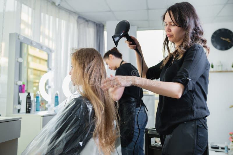 Cabeleireiro secando cabelos loiros à mão com secador de cabelo e escova  redonda em um salão de beleza.