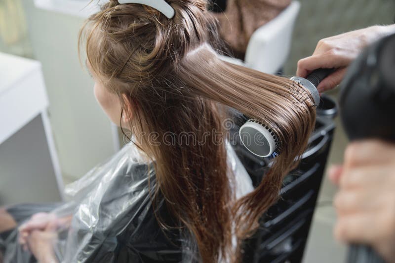 Cabeleireiro secando cabelos loiros à mão com secador de cabelo e escova  redonda em um salão de beleza.