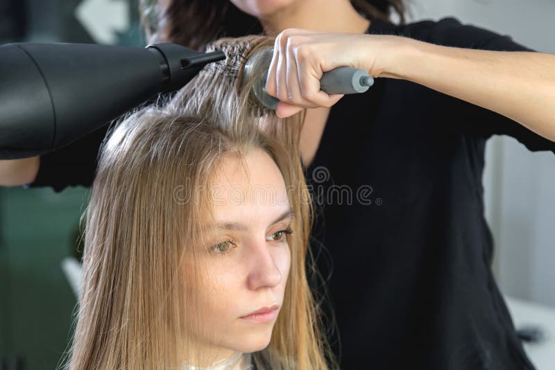 Cabeleireiro secando cabelos loiros à mão com secador de cabelo e escova  redonda em um salão de beleza.