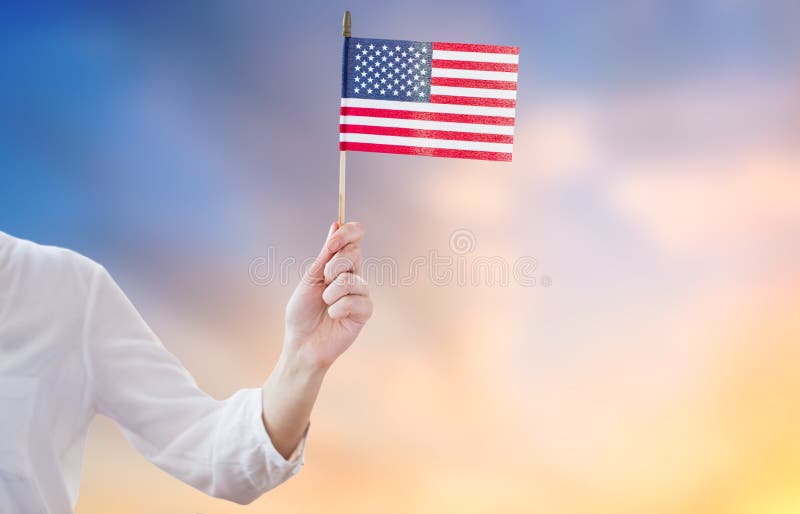 Independence day, celebration, patriotism and holidays concept - close up of woman holding american flag in hand at 4th july party over evening sky background. Independence day, celebration, patriotism and holidays concept - close up of woman holding american flag in hand at 4th july party over evening sky background