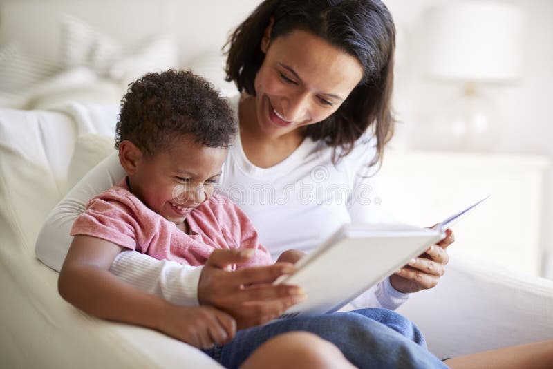 Feche acima da mãe adulta nova afro-americano que senta-se em uma poltrona que lê um livro com seu filho da criança de três anos