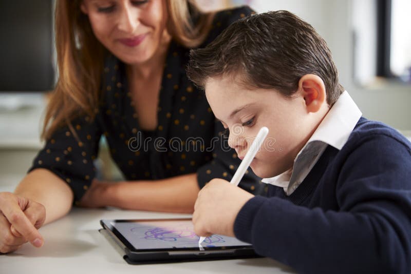 Feche acima do professor fêmea novo que senta-se na mesa com uma estudante de Síndrome de Down que usa um tablet pc em um classr