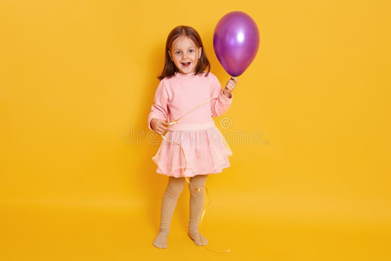 Feche o retrato de uma garotinha fofa segurando um balão de puxo e parece expelida colocando-se isolada sobre o fundo amarelo, en