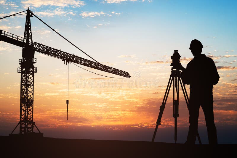 Silhouette Of A Surveyor Standing With Equipment Near Crane At Construction Site During Sunset. Silhouette Of A Surveyor Standing With Equipment Near Crane At Construction Site During Sunset