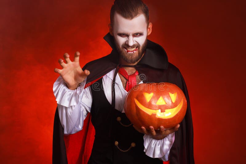 Felice Halloween! uomo barbuto in un costume da vampiro di Dracula con una zucca su uno sfondo rosso brillante fotografia stock Felice Halloween! uomo barbuto in un costume da vampiro di Dracula con una zucca su uno sfondo rosso brillante fotografia stock