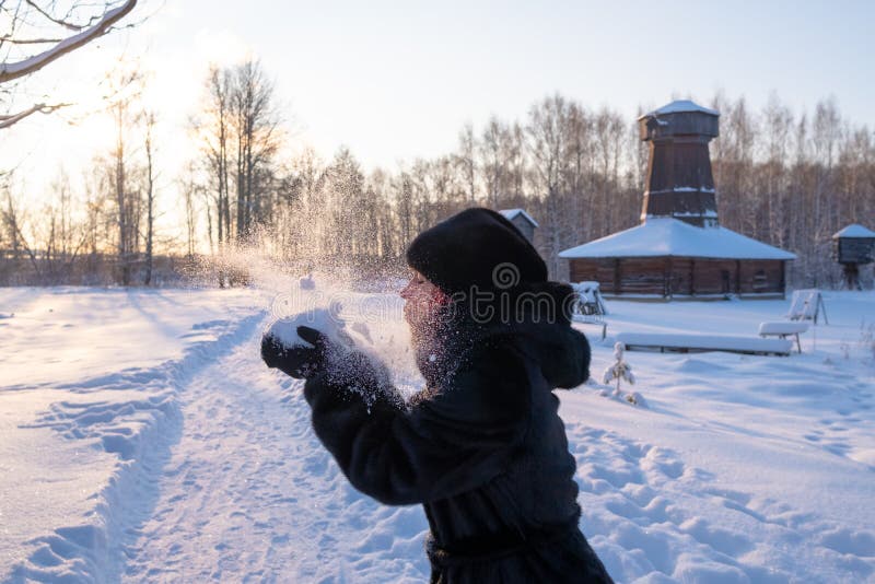 Felice ragazza con una pelliccia nera e un cappello di visone le fa saltare la neve dalle mani Al tramonto fiocchi di neve luccic fotografia stock Felice ragazza con una pelliccia nera e un cappello di visone le fa saltare la neve dalle mani Al tramonto fiocchi di neve luccic fotografia stock