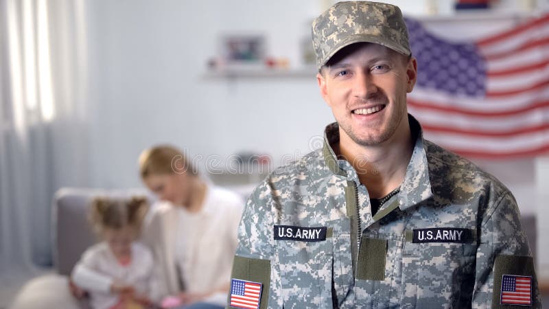Felice soldato dell'esercito USA che sorride alla macchina fotografica, moglie e figlia seduta sul divano