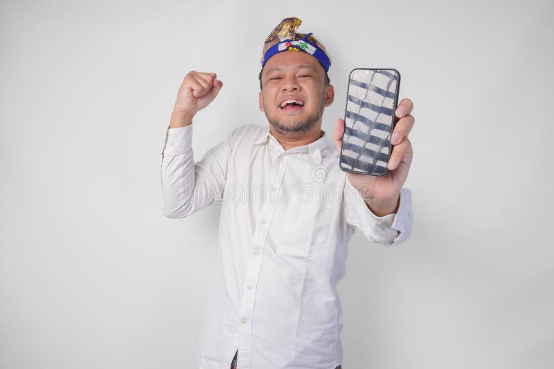 Felicissimo giovane balinese con camicia bianca e copricapo tradizionale presentando uno schermo vuoto, copia lo spazio sul telefo immagine stock libera da diritti